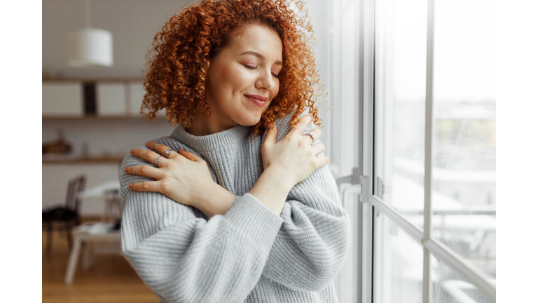 Horizontal image of pretty redhead female with closed eyes wearing rings on fingers, hugging herself, touching new soft sweater, enjoying comfort of fabric, standing next to panoramic window at home