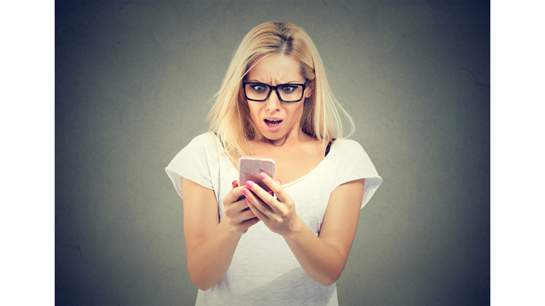 Shocking message. Surprised woman holding mobile phone and staring at it while standing against gray background