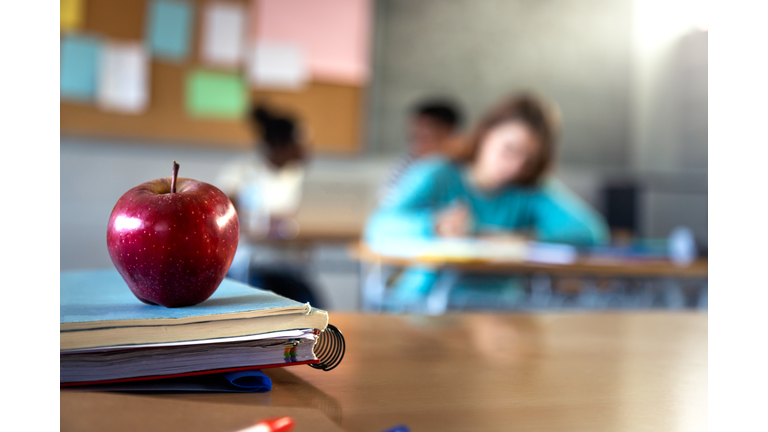 Apple and a pile of books and pens on teacher table in classroom. Focus on foreground.