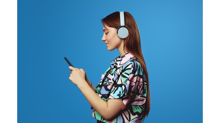 Side view of young student girl holding mobile phone and wearing headphones while listening to music