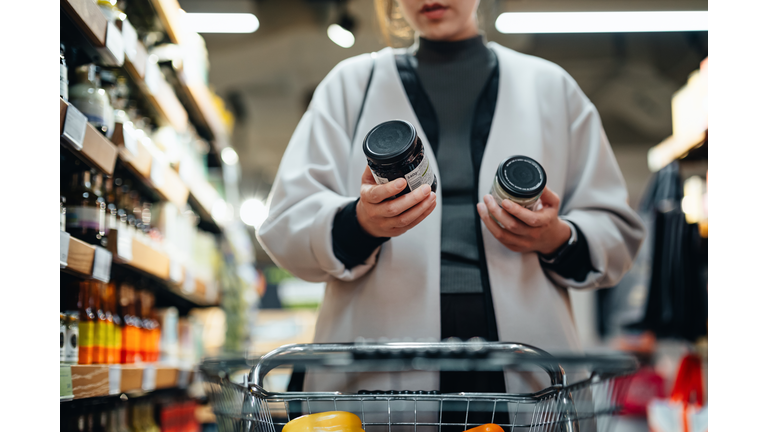 Young Asian woman with shopping cart shopping for cooking sauce in supermarket, reading and comparing the product information of two bottles of cooking sauces. Routine grocery shopping. Making healthier food choices. Consumer awareness concept