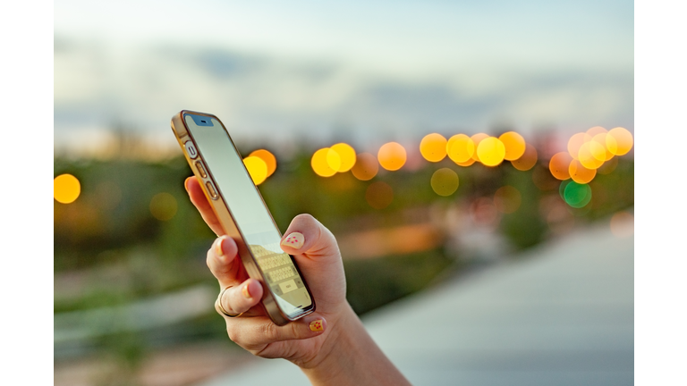 Young woman's hand holding and using her iPhone against a twilight urban background.