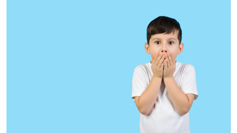 Surprised child in a white t-shirt on studio blue isolated background. Boy covers his mouth with his hands