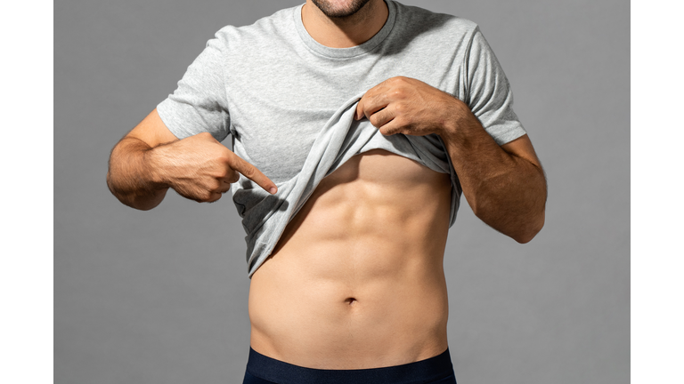 Muscular male model lifting up t-shirt to show abs while standing on gray isolated background in studio