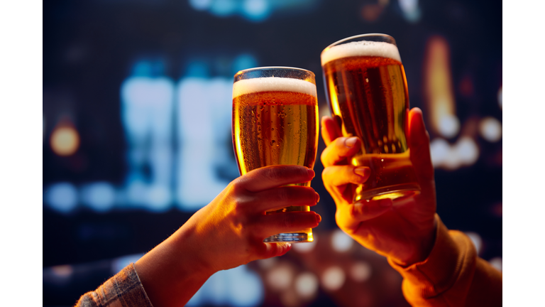 Hands holding lager light foamy big beer glasses and clinking. Two friends sitting in bar and drinking alcohol drinks.