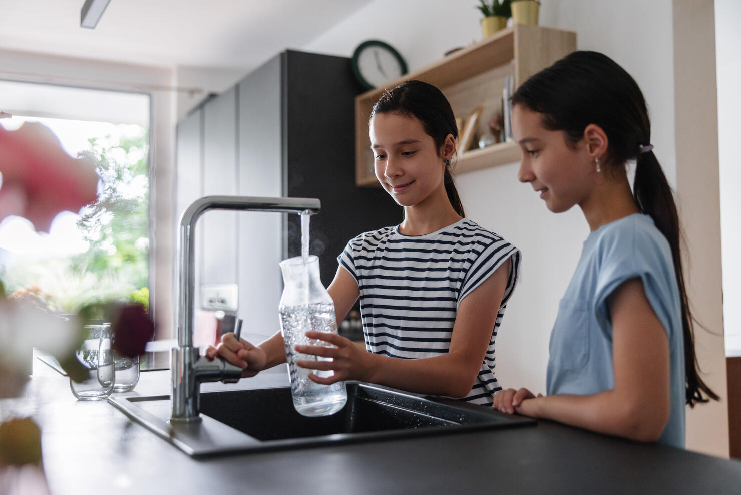 Side View Of Two Young Girls At The Kitchen Filling A Glass Jar With Some Tap Water