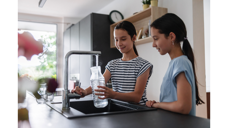 Side View Of Two Young Girls At The Kitchen Filling A Glass Jar With Some Tap Water