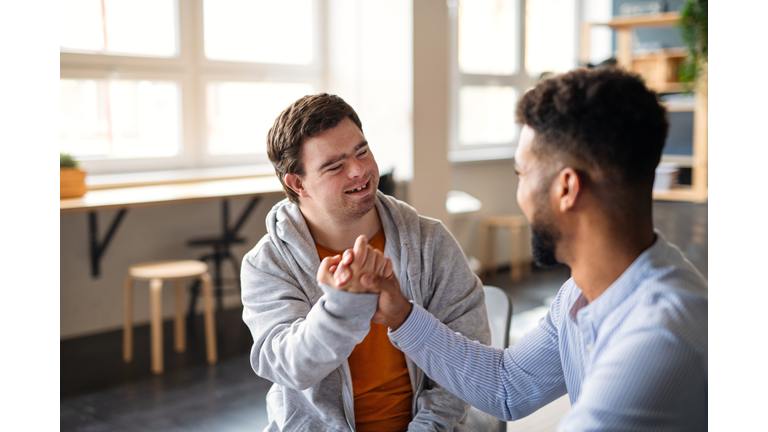 Young happy man with Down syndrome with his mentoring friend celebrating success indoors at school.