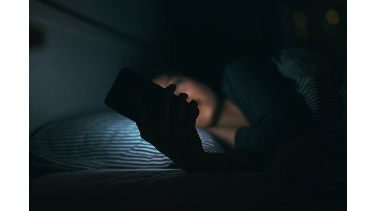 Young woman text messaging on smartphone while relaxing and lying on bed at night
