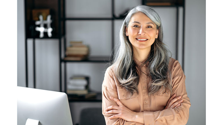 Portrait of a successful confident mature gray-haired lady, business woman, ceo or business tutor, standing in the office with arms crossed, looking and friendly smiling into the camera