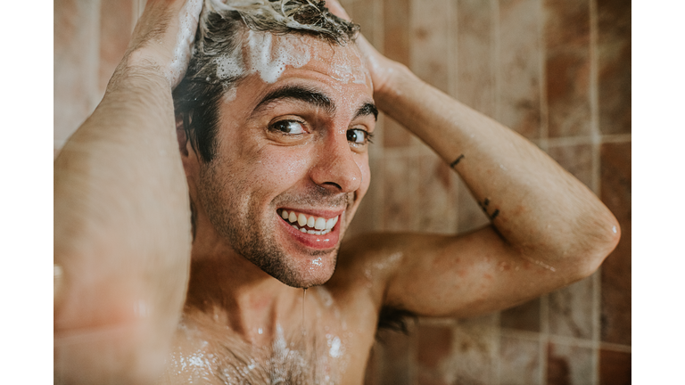 A man washes his hair in the shower. He is confident and happy as he smiles into the camera.