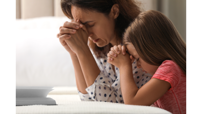 Mature woman with her little granddaughter praying together over Bible in bedroom