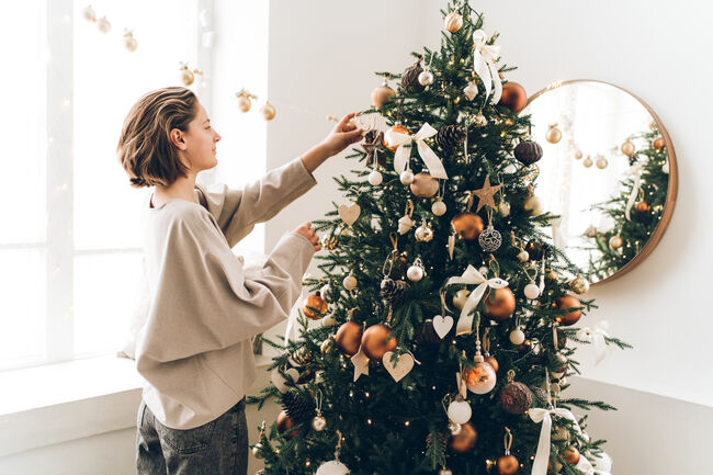 Young woman is adjusting her hair and decorating Christmas tree with many different decorations and festive garland. New Year celebration concept