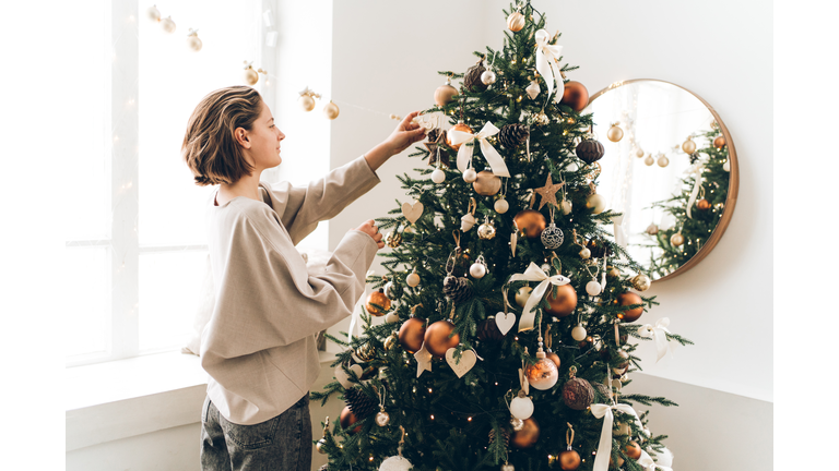 Young woman is adjusting her hair and decorating Christmas tree with many different decorations and festive garland. New Year celebration concept