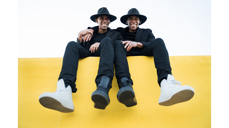 Smiling African American twin brothers wearing black funny clothes and hat embracing each other, sitting on yellow wall