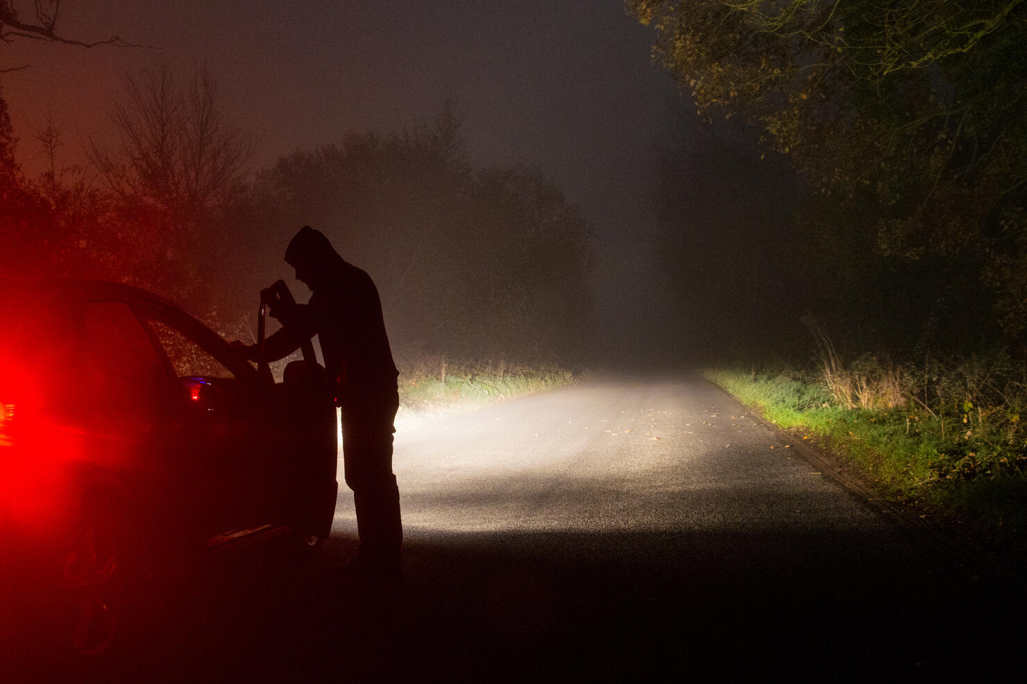 A sinister hooded figure opening a car door, on a empty country road at night