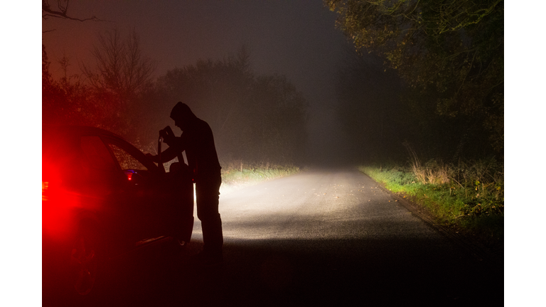 A sinister hooded figure opening a car door, on a empty country road at night