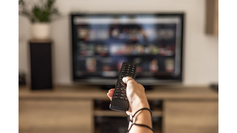 A woman is holding a remote control of a smart TV in his hand. In the background you can see the television screen with streaming entertainment apps for video on demand.