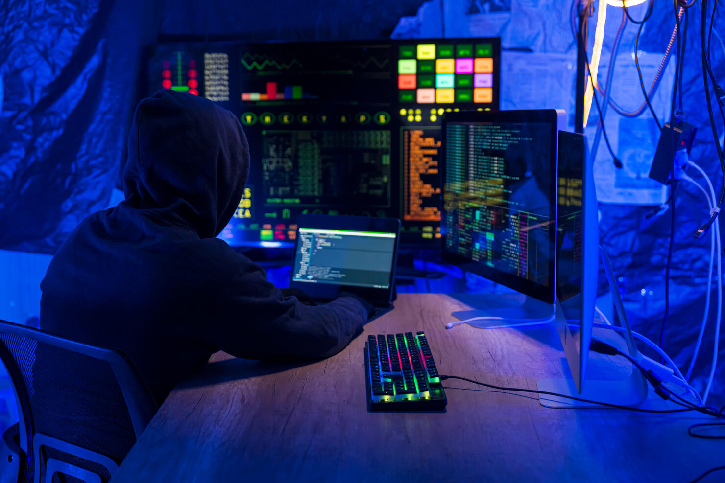 Rear view of computer hacker in dark hall wearing hooded top hacks into a computer network, types software code on a keyboard and monitors a virus attack to hack into network systems and servers.