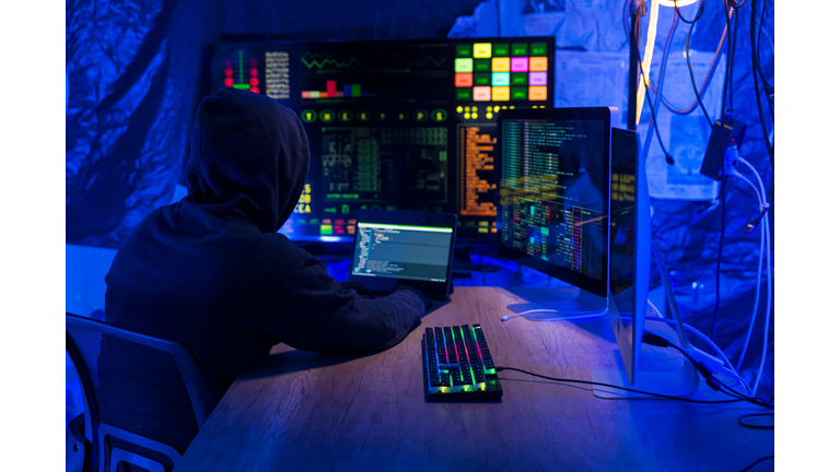Rear view of computer hacker in dark hall wearing hooded top hacks into a computer network, types software code on a keyboard and monitors a virus attack to hack into network systems and servers.