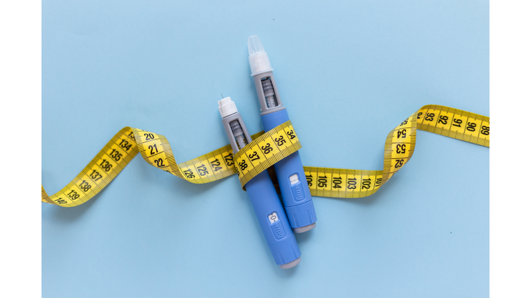 Two injectors dosing pens for subcutaneous injection of antidiabetic medication or anti-obesity medication hovering over a blue background. Yellow measuring tape around the injectors.