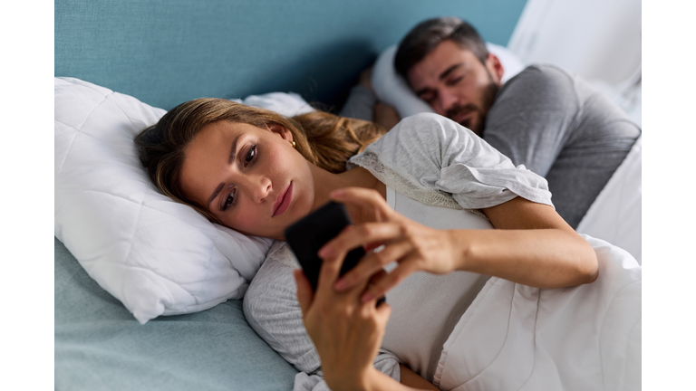 Young woman using mobile phone and checking messages in bed while her husband is asleep