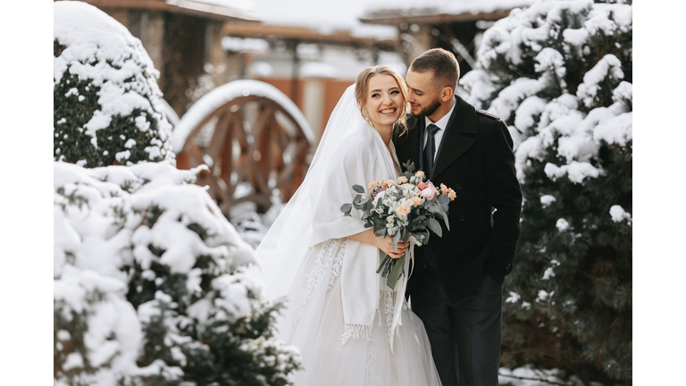 Beautiful and happy bride and groom are having fun among snowy trees. Bride and groom in the winter park. Bride with a bouquet of flowers in a wedding dress and poncho. Groom in a black coat.