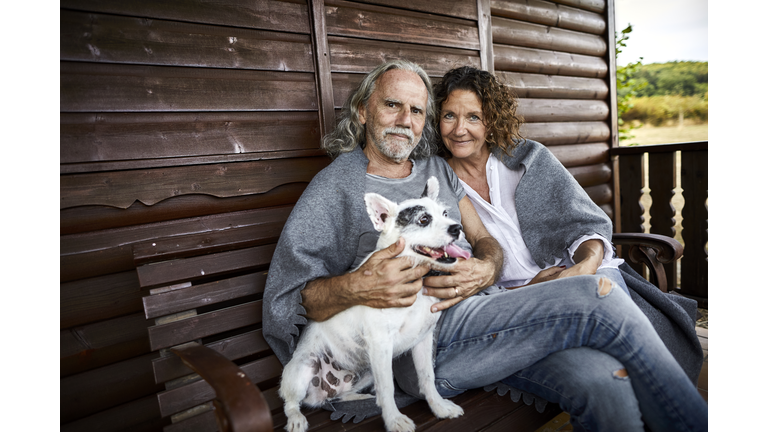 Portrait of mature couple with dog sitting on porch of a log cabin