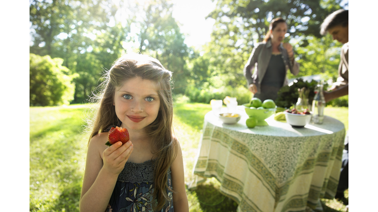 Outdoors in summer. On the farm. Children and adults together. A young girl holding a large fresh organically produced strawberry fruit. Two adults beside a round table.