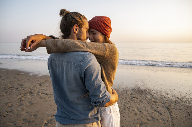 Young romantic couple standing face to face at beach against clear sky during sunset