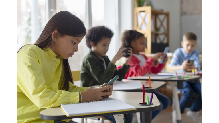 Diverse multiethnic kids students having break and using smartphones, sitting at desks in classroom, children playing games and checking social media at school