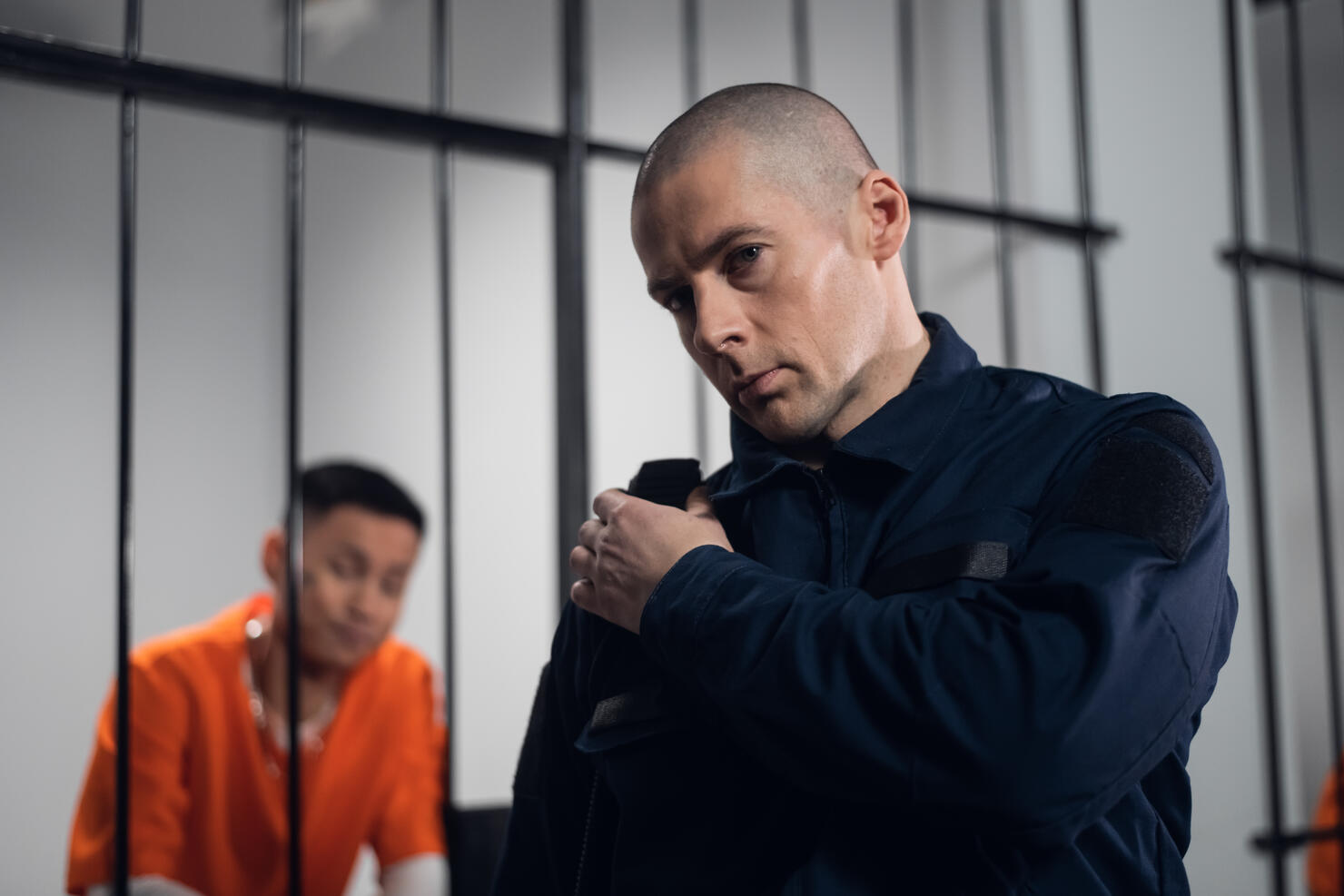 A prison guard in full ammunition communicates with his partner on the radio. In the background, a prisoner in a cell