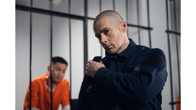 A prison guard in full ammunition communicates with his partner on the radio. In the background, a prisoner in a cell