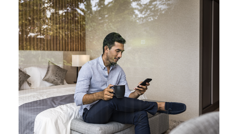 Young Asian businessman  working from home, using smartphone, sitting on bed at bedroom  window of modern house