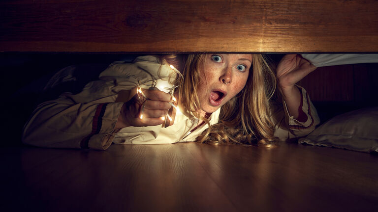 Young woman with emotional face, holding lights, lying on floor and looking under bed with emotions of fear. Spooky season