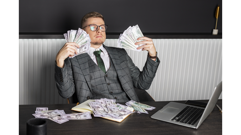 A young serious man in a gray formal suit and glasses sits at his desk and counts cash. A businessman holds money.