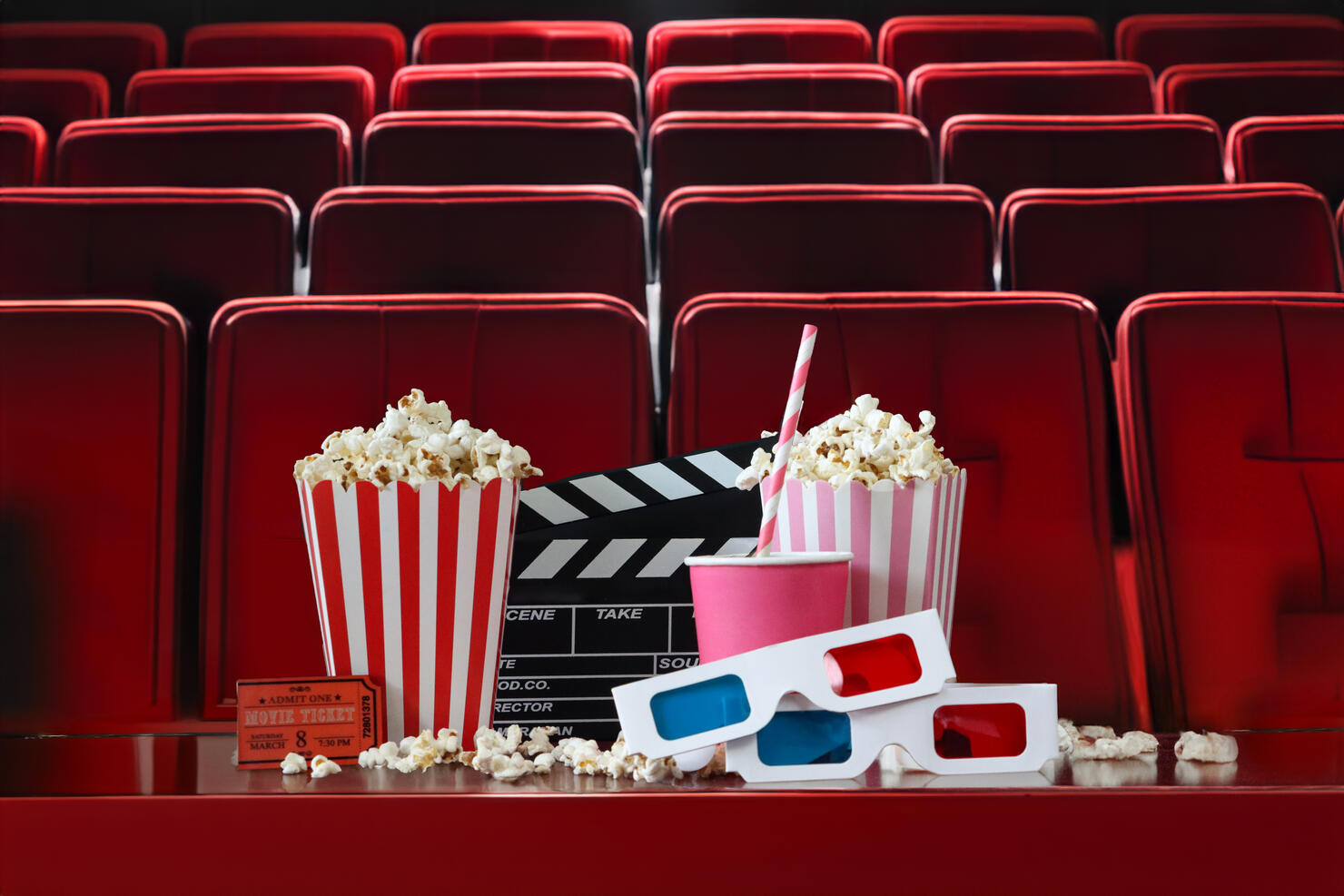 Close-up image two striped cartons of popcorn surrounded by spilled popped kernels, pink disposable drinks cup and drinking straw, two pairs of 3D glasses, cinema theatre ticket, black film clapperboard, open hinged clapper stick, cinema seats background