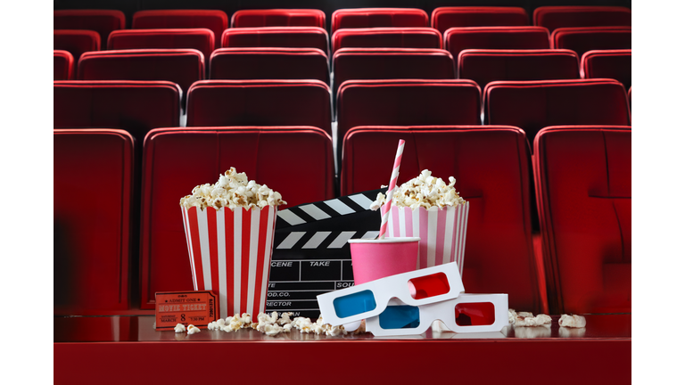 Close-up image two striped cartons of popcorn surrounded by spilled popped kernels, pink disposable drinks cup and drinking straw, two pairs of 3D glasses, cinema theatre ticket, black film clapperboard, open hinged clapper stick, cinema seats background