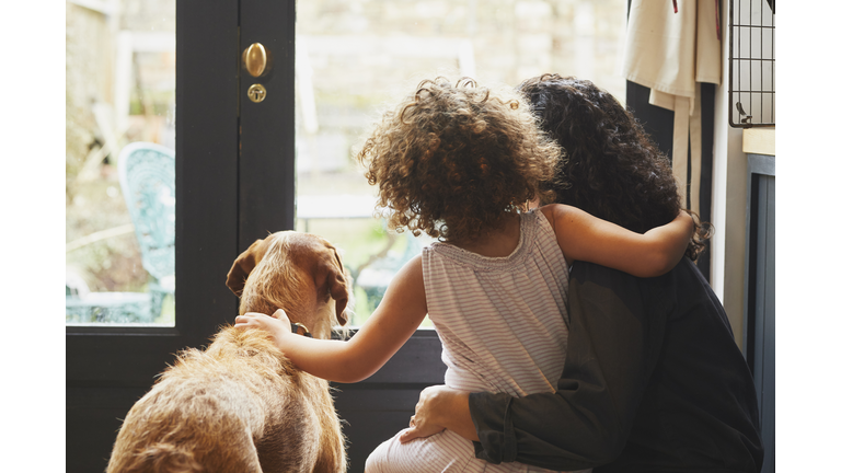 Back view of mixed race Mother and young daughter with dog looking out of kitchen window.