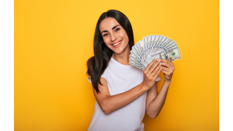 Young happy excited beautiful woman emotionally looking in the camera with a lot of money in her hands and having fun isolated on yellow background