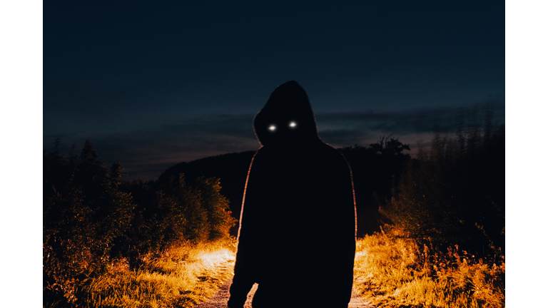 A horror concept of a scary hooded figure with glowing eyes. At night back lighted in the countryside