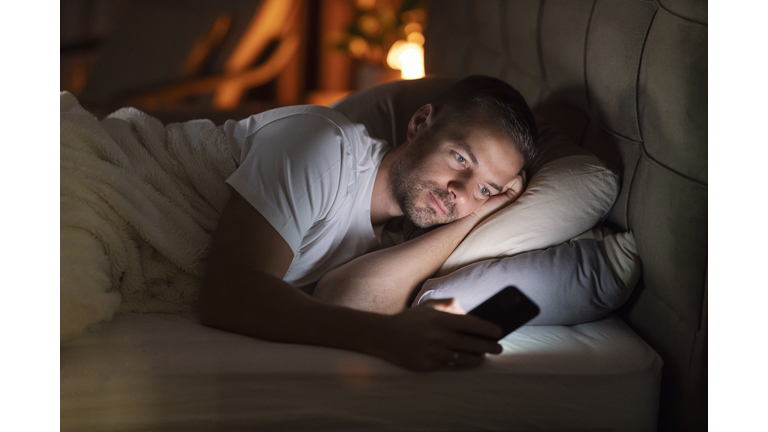 A sleepless man lying in dark bedroom with mobile phone in hands and scrolling endlessly.