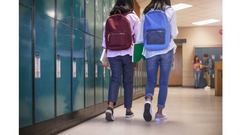 Female classmate friends with backpacks walking by lockers in school