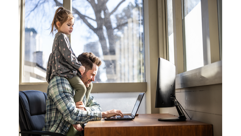 Father working in home office while toddler daughter sits on his shoulders