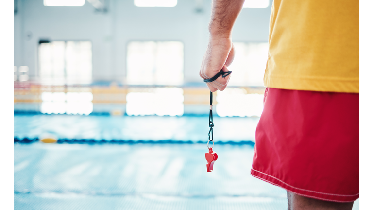 Hands, lifeguard and whistle by swimming pool for water safety, security or ready for rescue indoors. Hand of expert swimmer holding signal tool for warning, safe swim or responsibility for awareness