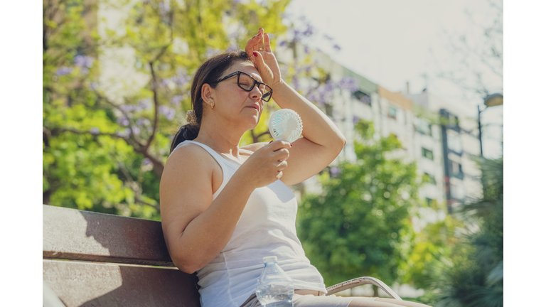 Woman suffering from heat stroke and headache. Holding an electric mini fan sitting outdoors