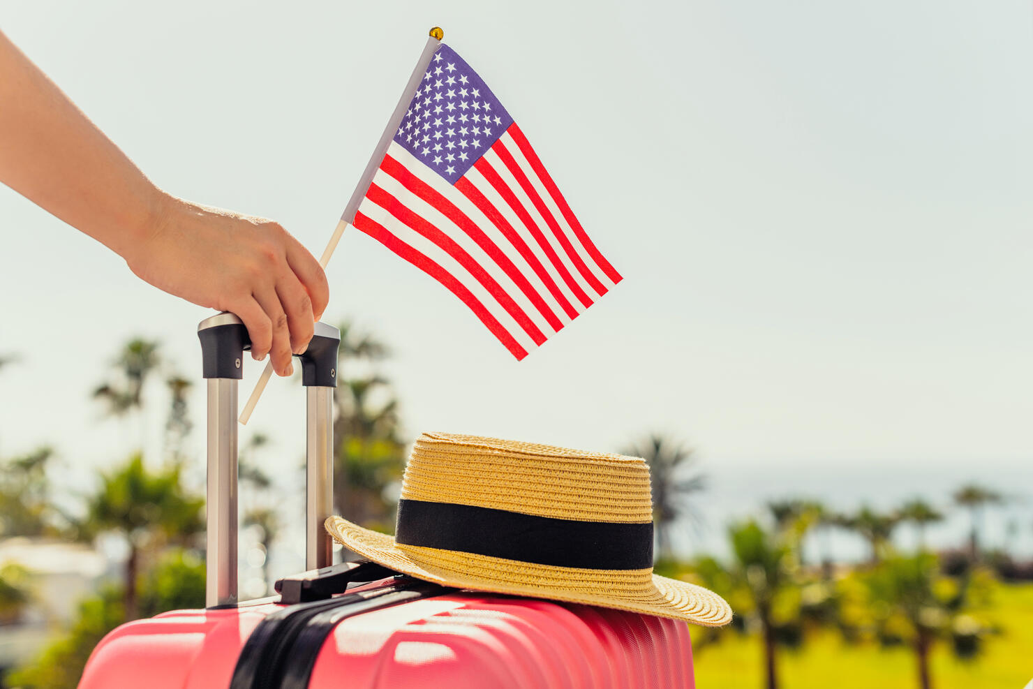 Woman with pink suitcase, hat and America flag standing on passengers ladder and getting out of airplane opposite sea coastline with palm trees.
