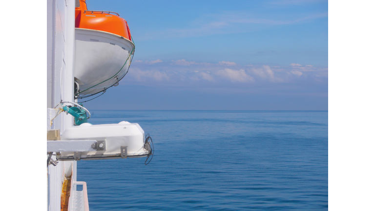 Lifeboat on the deck of a ferry cruise ship. Saving lives on the water concept. Side view against the background of the sky and calm sea in sunny weather.