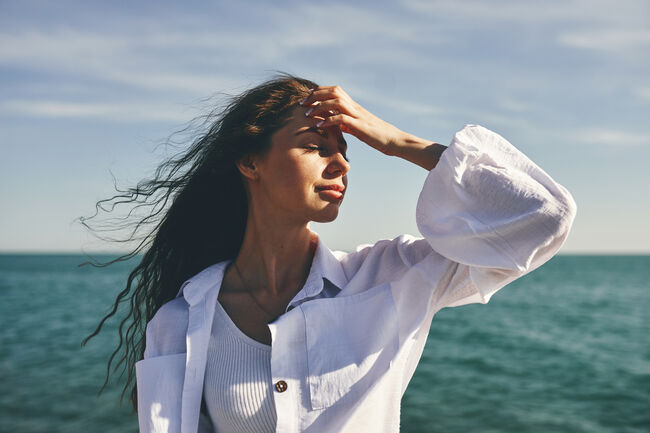 woman standing by the sea with hand on forehead. Hair blowing in the wind. Clear sky