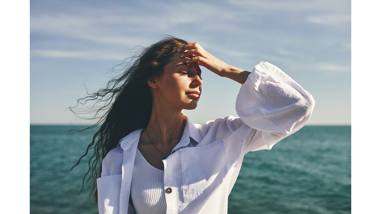 woman standing by the sea with hand on forehead. Hair blowing in the wind. Clear sky